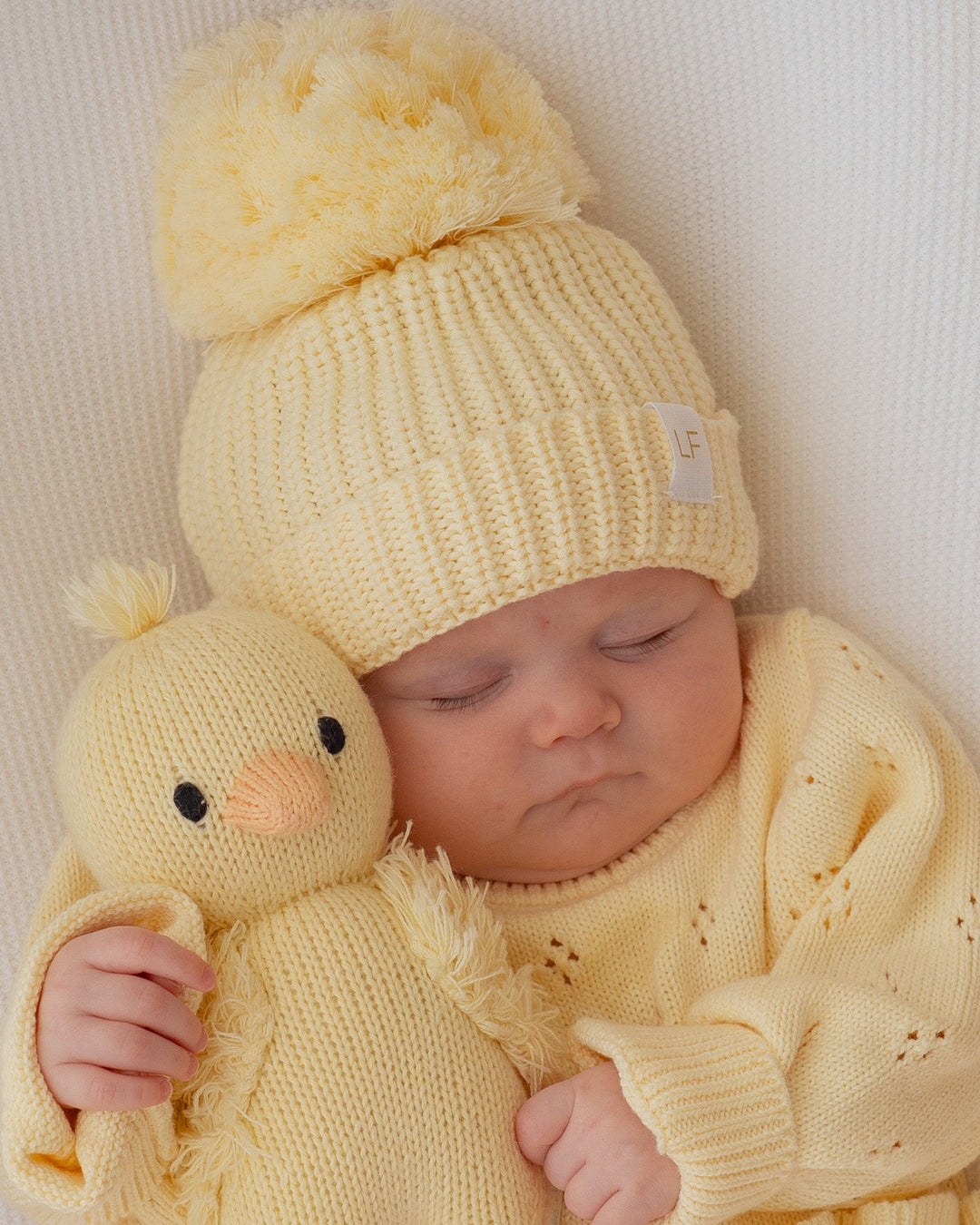 Baby in yellow outfit holding a yellow knitted duck toy on a pink blanket with 'Stella' printed on it.