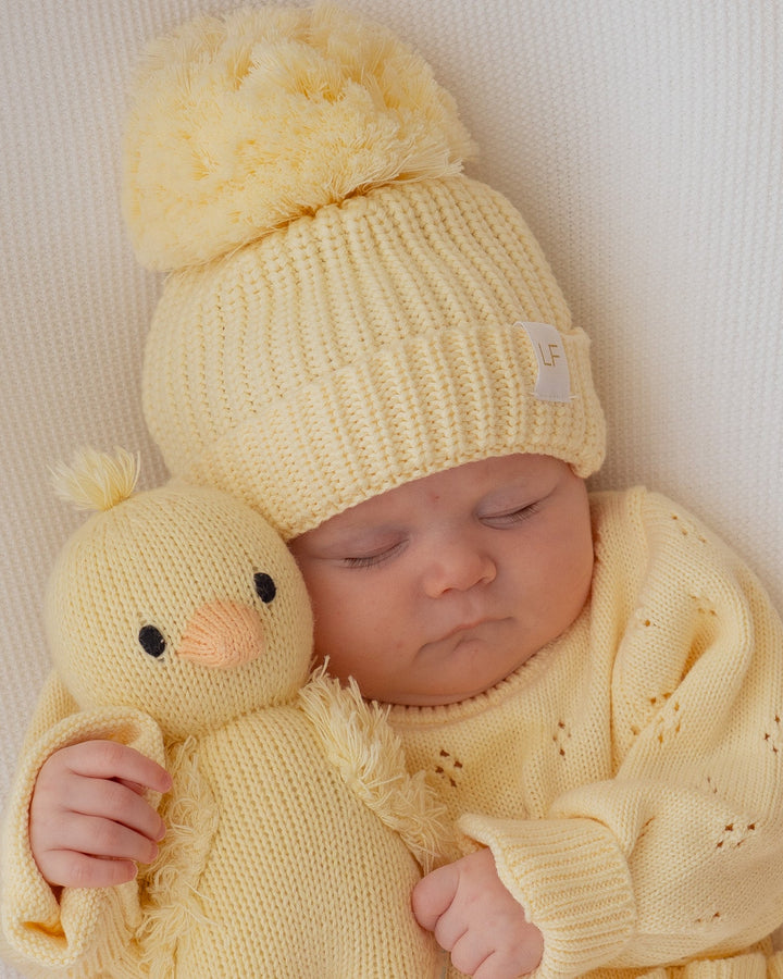 Baby in yellow outfit holding a yellow knitted duck toy on a pink blanket with 'Stella' printed on it.