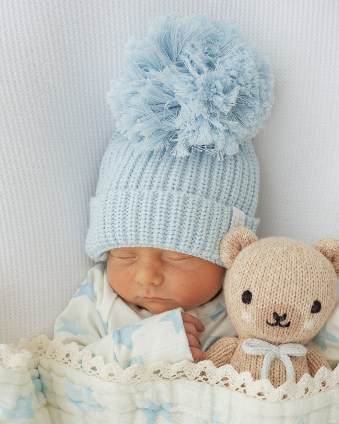 Newborn baby wrapped in a blanket with a knitted blue hat and holding a teddy bear.