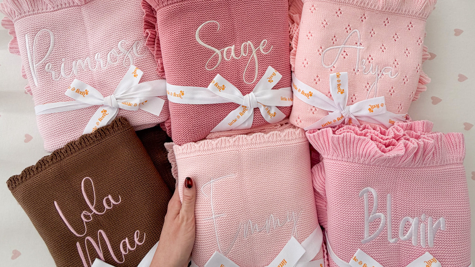 Six personalized baby blankets in pink, dusty pink, and brown are arranged in a crib. Each blanket has a ruffled edge, custom script embroidery, and a white ribbon tied in a bow with a 'Belle' print.