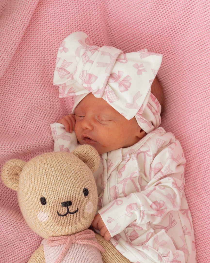 Newborn baby wrapped in pink blanket with teddy bear and bow headband on pink background